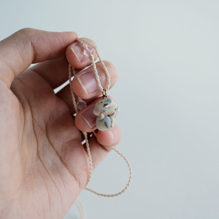A hand holding a delicate necklace with a small, light gray and white textured pendant on a thin beige cord, set against a plain light gray background.